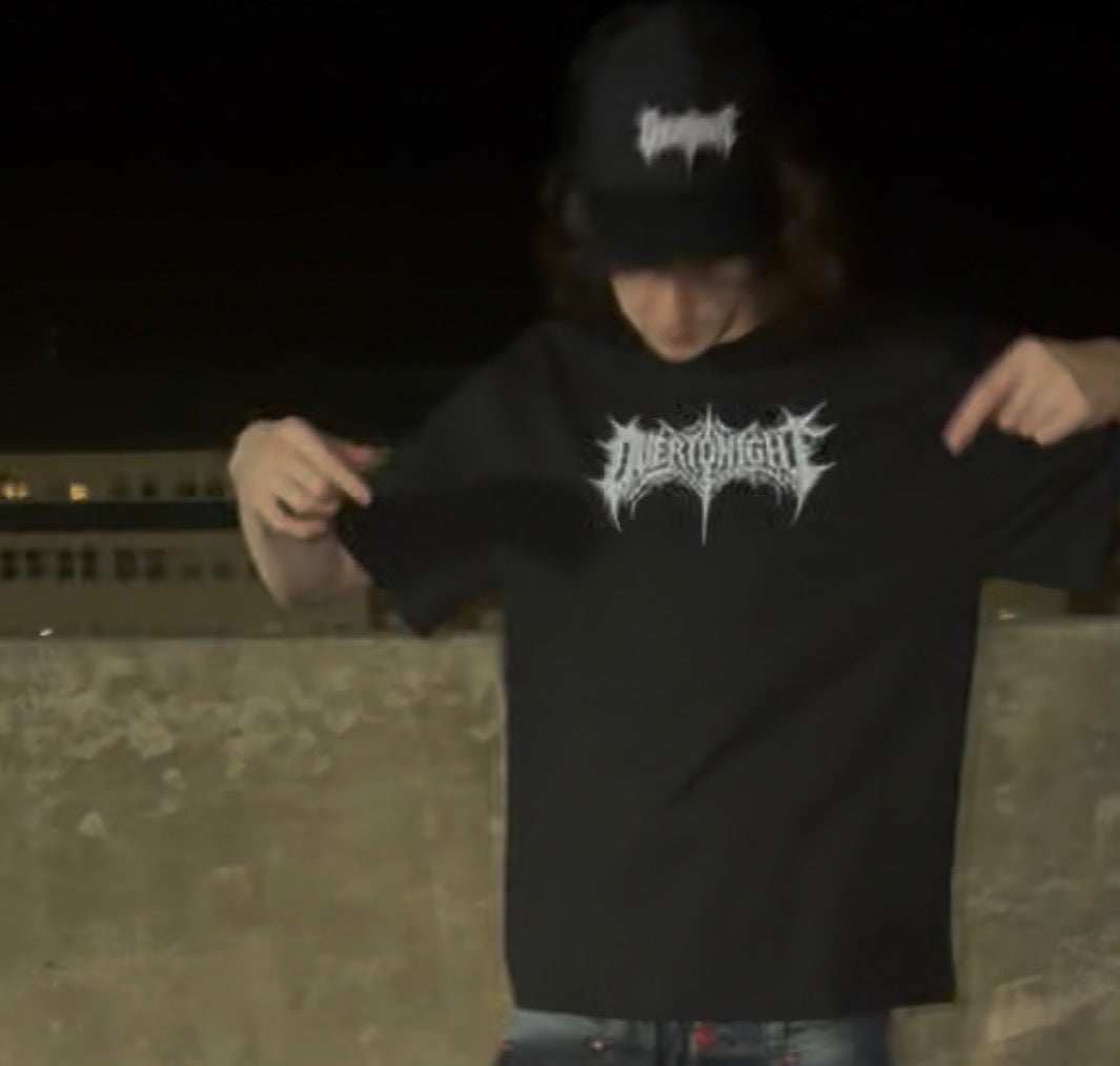 A person at night wearing a black t-shirt with a white overtonight logo and a matching black cap, pointing to the chest graphic.