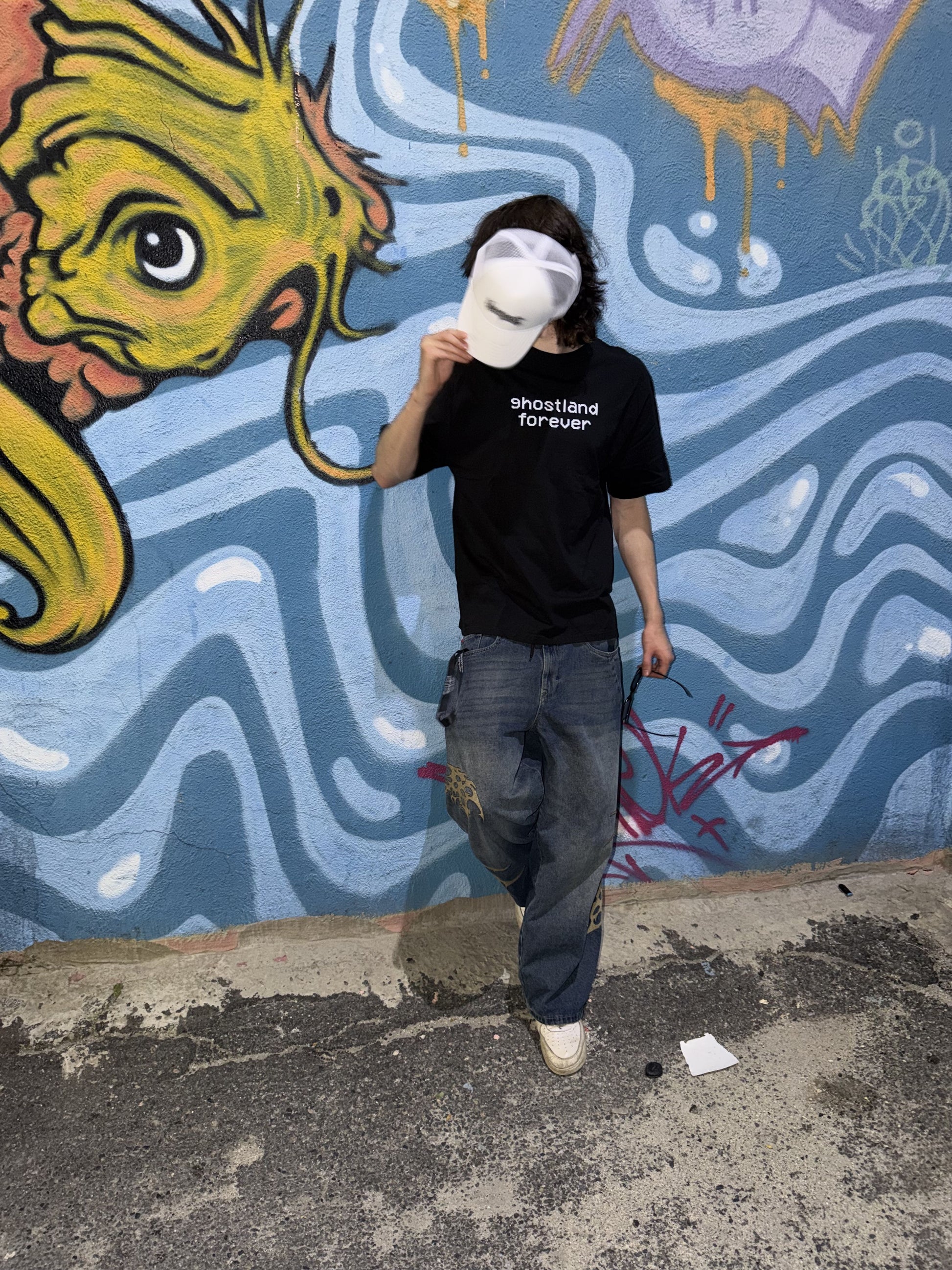 A person stands in front of a colorful graffiti wall wearing a black t-shirt with 'ghostland forever' printed in white, blue jeans, and a white visor.