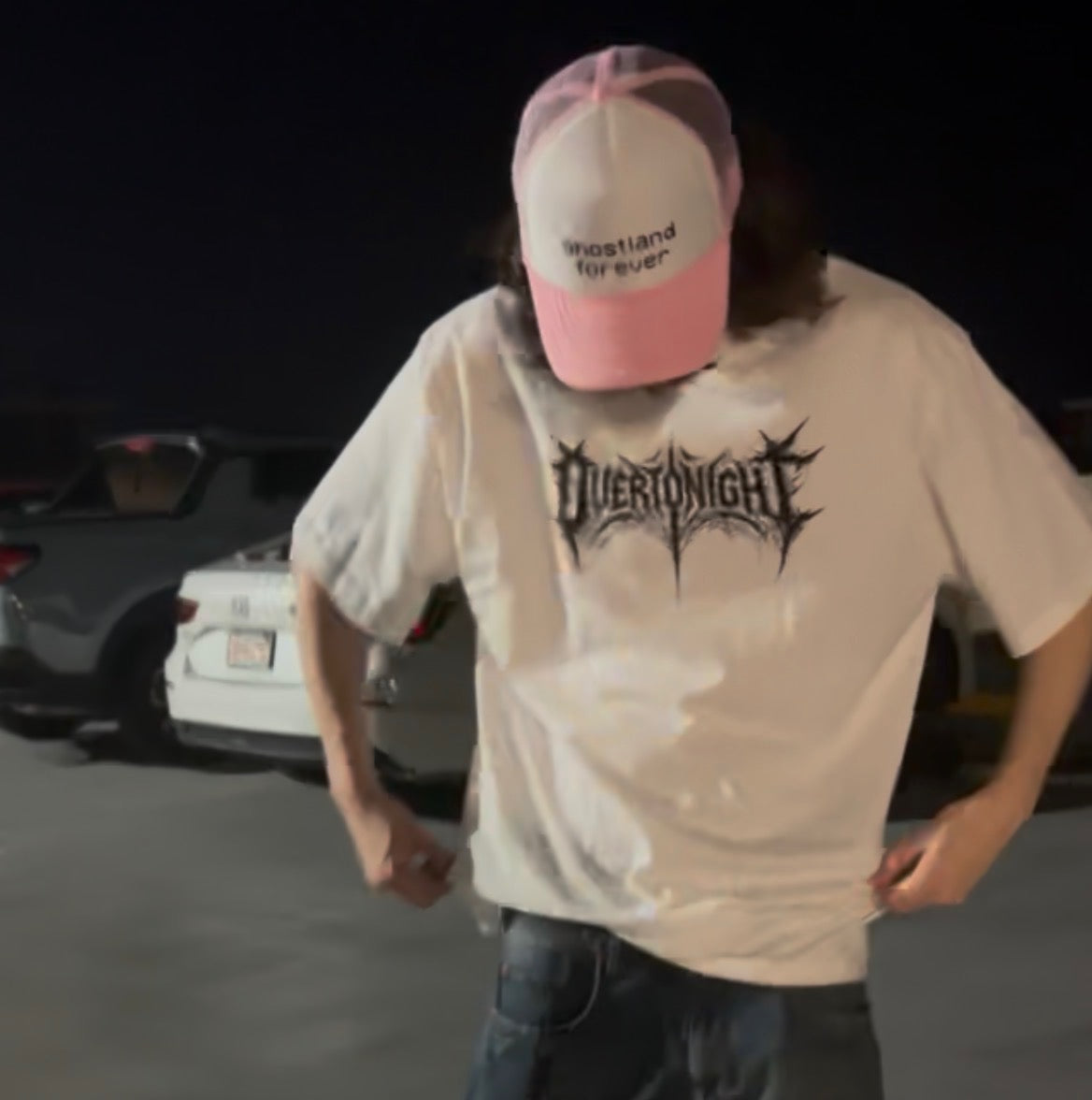 A person wearing an oversized white graphic t-shirt with a black 'Overtonight' band logo, paired with a pink and white trucker hat that says 'ghostland forever', standing in a dark outdoor parking area.