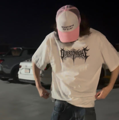 A person wearing an oversized white graphic t-shirt with a black 'Overtonight' band logo, paired with a pink and white trucker hat that says 'ghostland forever', standing in a dark outdoor parking area.