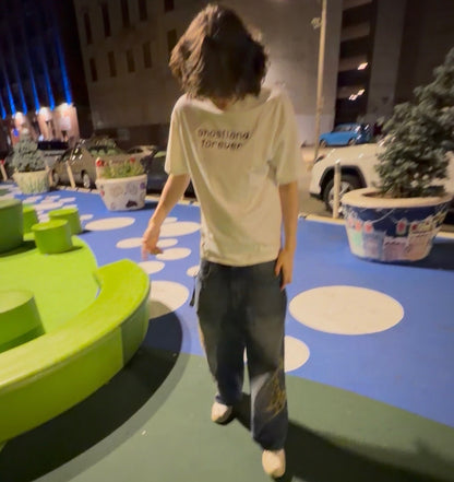 A person wearing a cream-colored graphic t-shirt with 'ghostland forever' printed on the back, paired with dark gray jeans and white shoes, standing in an outdoor urban plaza at night.