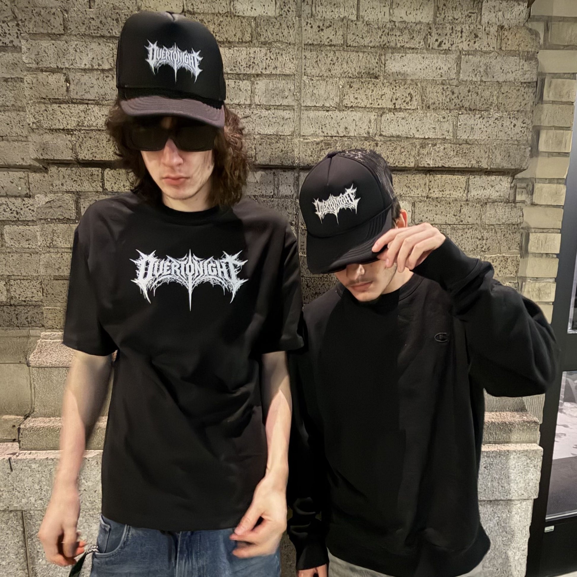 Two guys wearing matching black t-shirts and black baseball caps with a white gothic-style logo that reads 'overtonight'. One guy wears a black crewneck t-shirt and jeans, while the other wears a black long-sleeve hoodie.