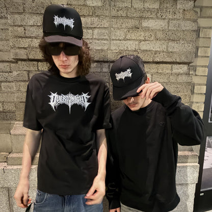 Two guys wearing matching black t-shirts and black baseball caps with a white gothic-style logo that reads 'overtonight'. One guy wears a black crewneck t-shirt and jeans, while the other wears a black long-sleeve hoodie.
