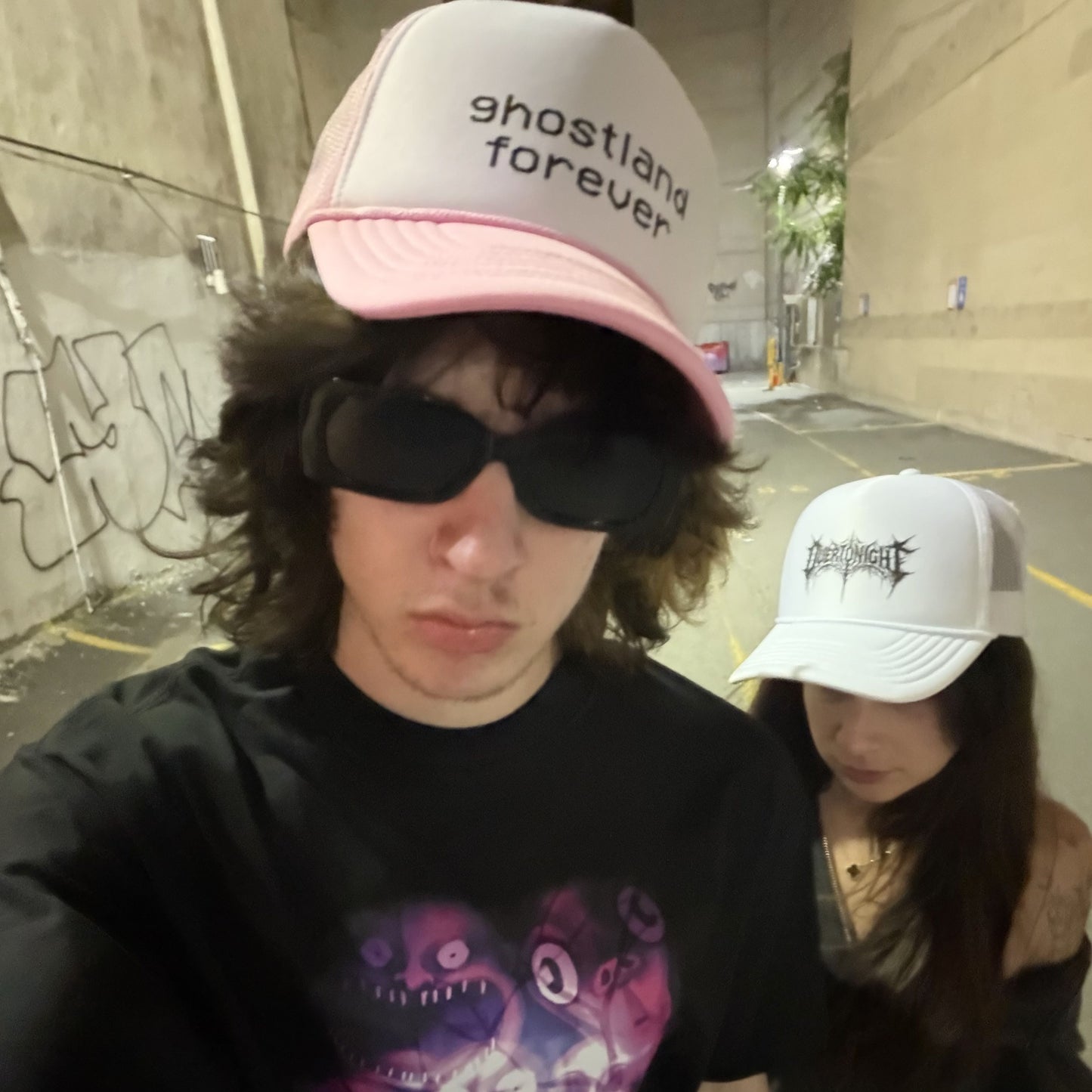 Two people taking a selfie in an outdoor concrete alley. The person in the foreground wears a pink and white trucker hat with the text 'ghostland forever' and a black graphic t-shirt.