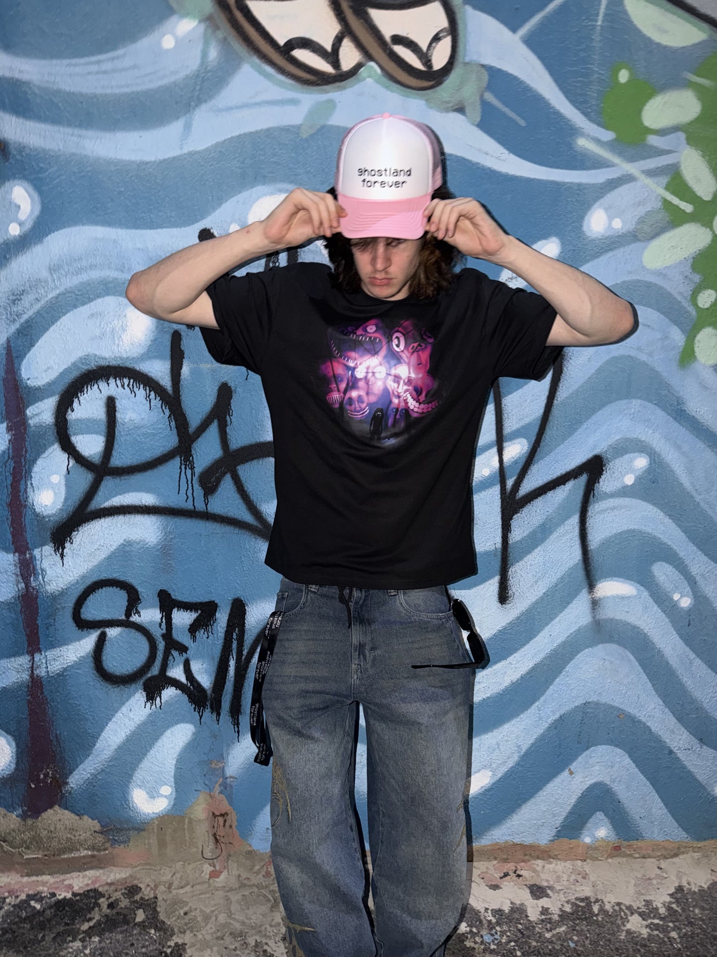 A person wearing a black graphic t-shirt, blue jeans, and a pink trucker hat with the tour design printed on the front, posing in front of a blue graffiti wall.