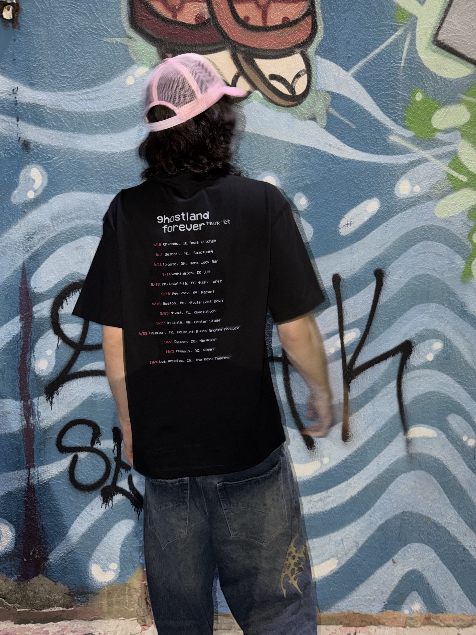 A person standing with their back to the camera, wearing a black t-shirt with tour dates on the back, blue jeans, and a pink and black trucker hat, set against a blue graffiti wall.