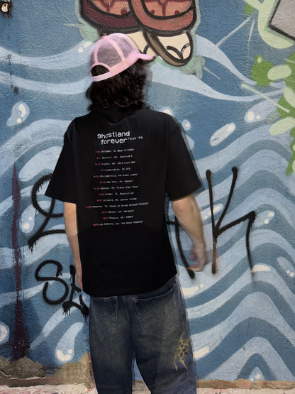 A person standing with their back to the camera, wearing a black t-shirt with tour dates on the back, blue jeans, and a pink and black trucker hat, set against a blue graffiti wall.